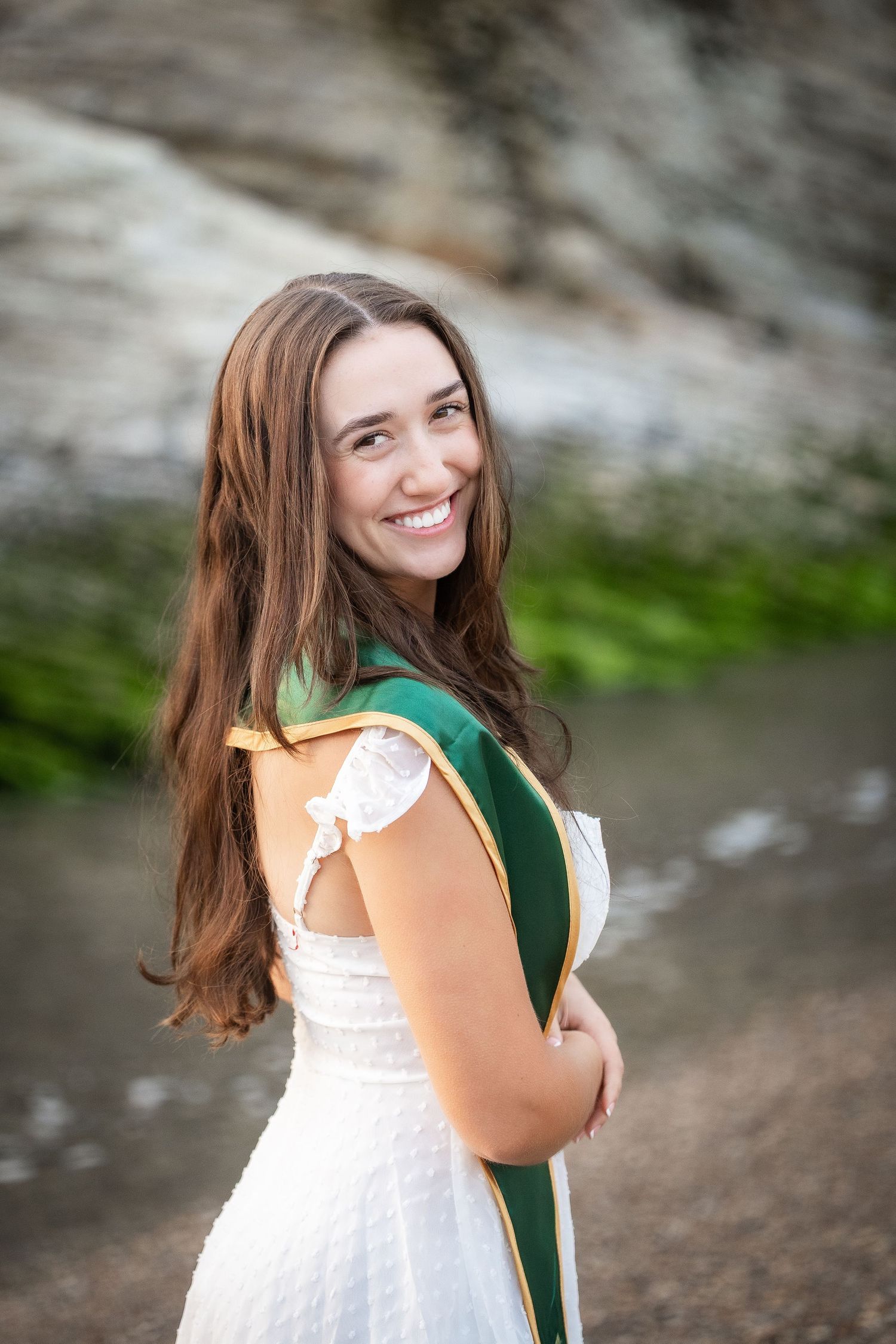 Woman in white dress and dark green scarf twirls joyfully outdoors against stone background.