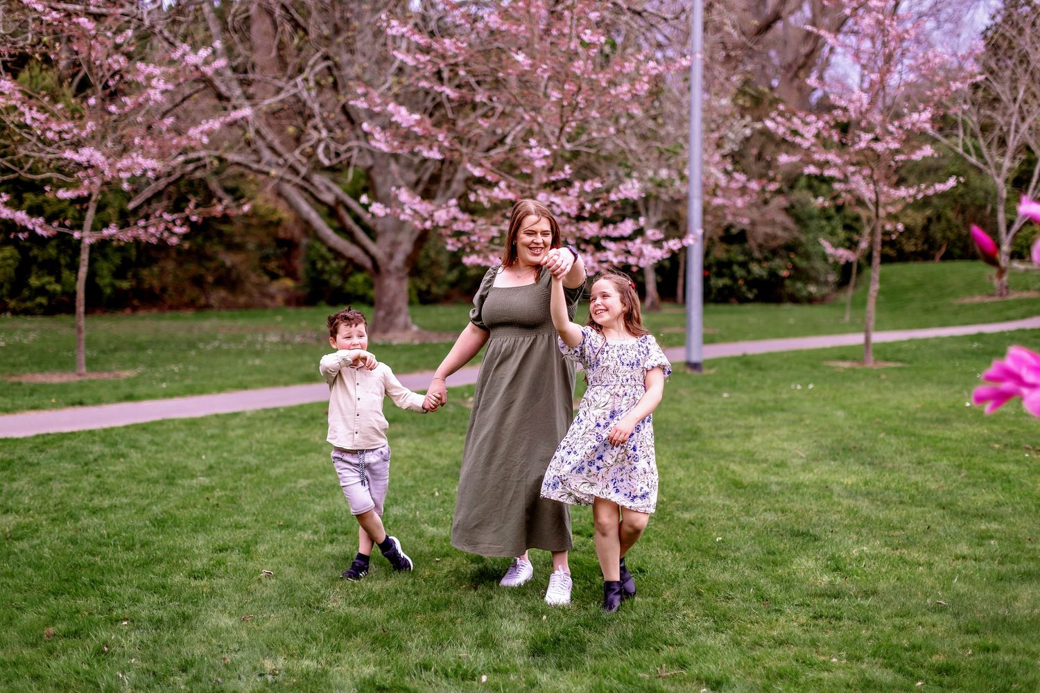 A mum and her children running and playing together beneath blooming pink cherry blossom trees in Bledisloe Park, Palmerston North.