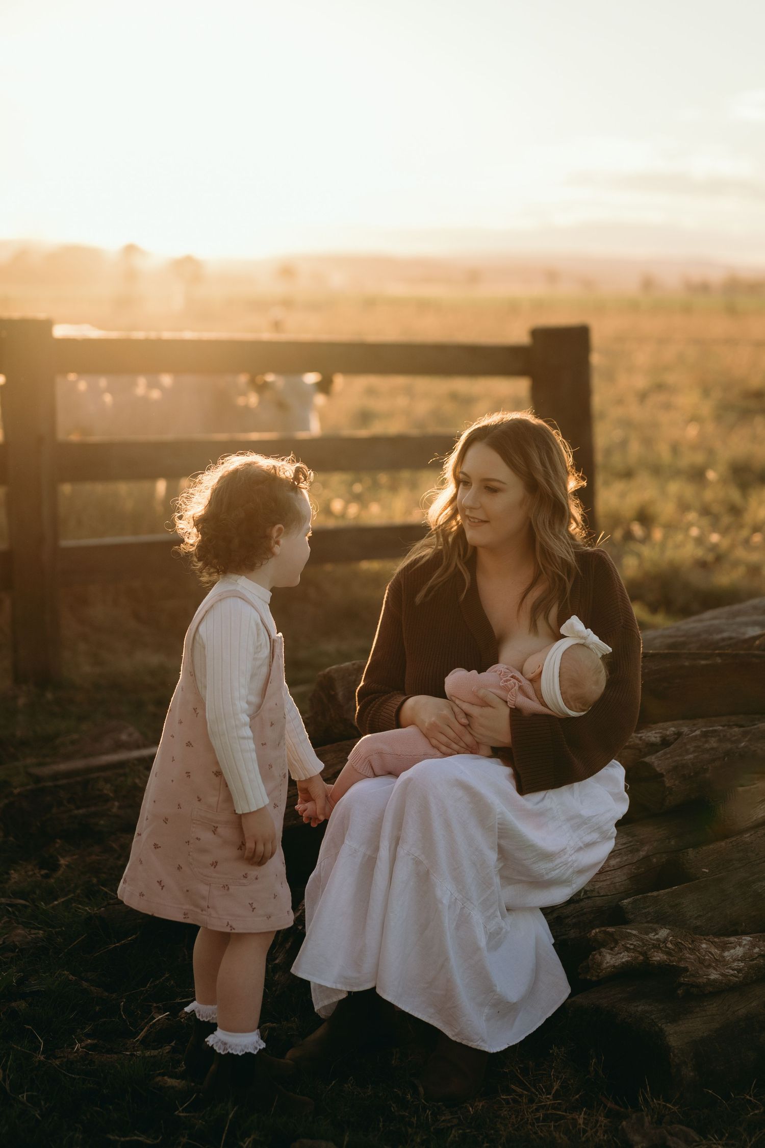 Mother with baby and young child sitting on fence during golden hour sunset.