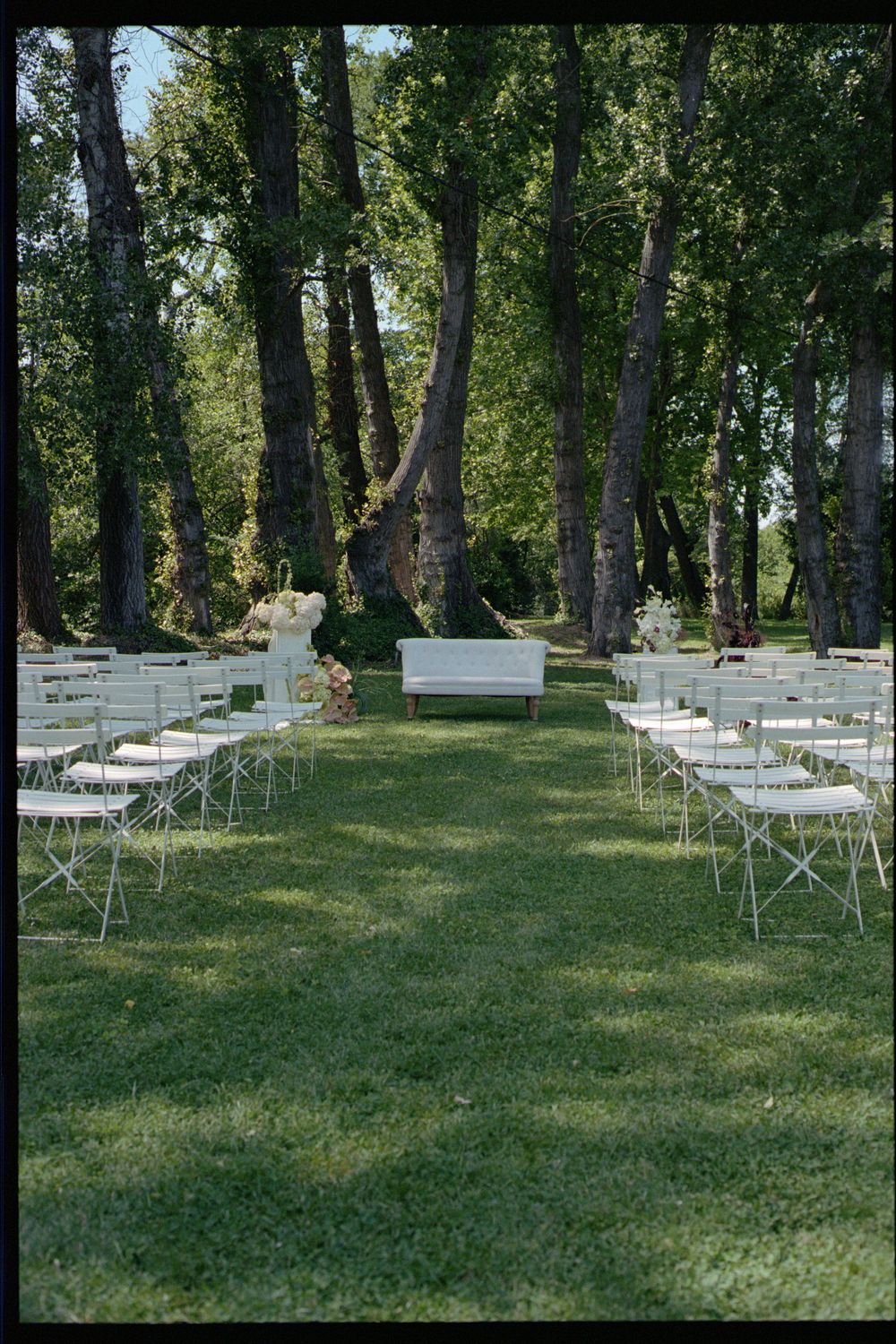 White folding chairs arranged on a grassy lawn surrounded by tall trees for an outdoor event or ceremony.