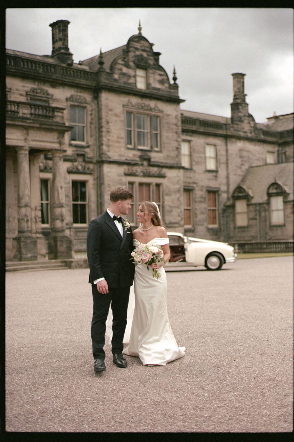 A wedding couple stands outside a grand stone mansion with Gothic architectural details and a vintage film aesthetic.