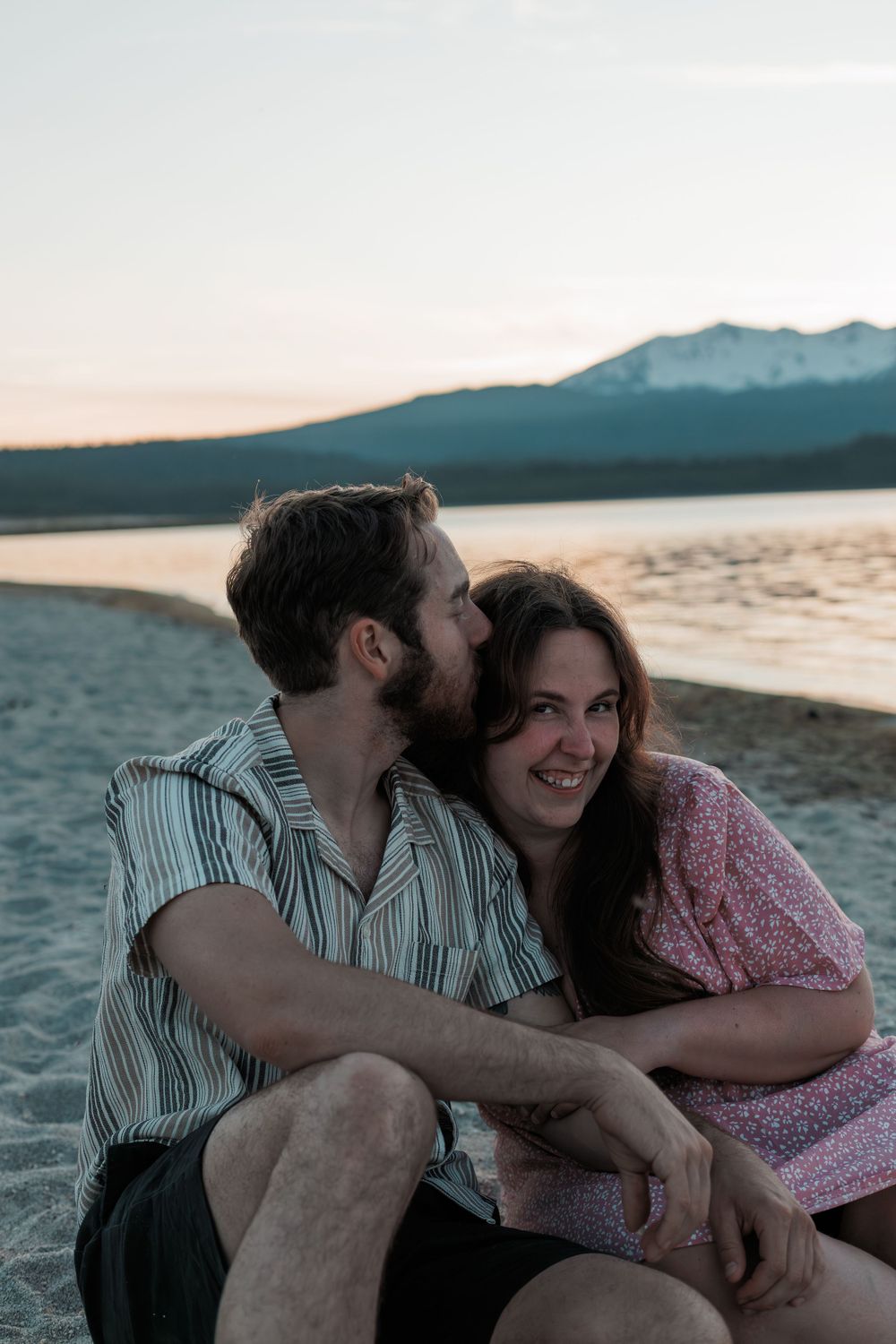 Couple sitting close together on a beach at sunset in Central Oregon with mountains in the background.