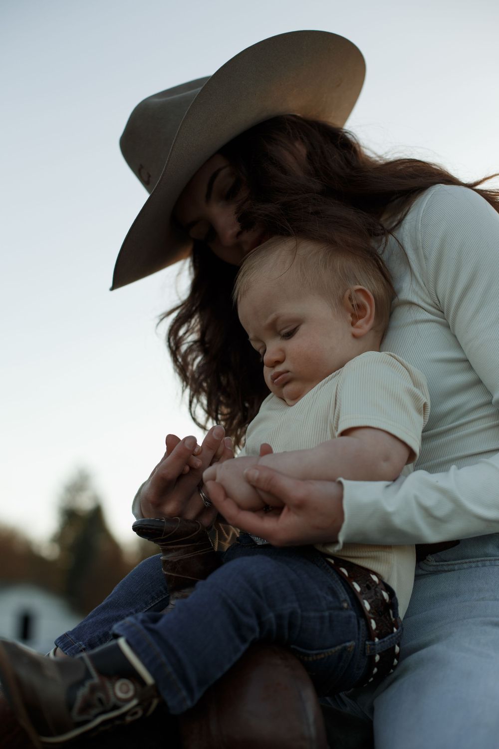 A tender moment captured outdoors at dusk as a mom in a cowboy hat holds her baby wearing a white shirt and jeans.