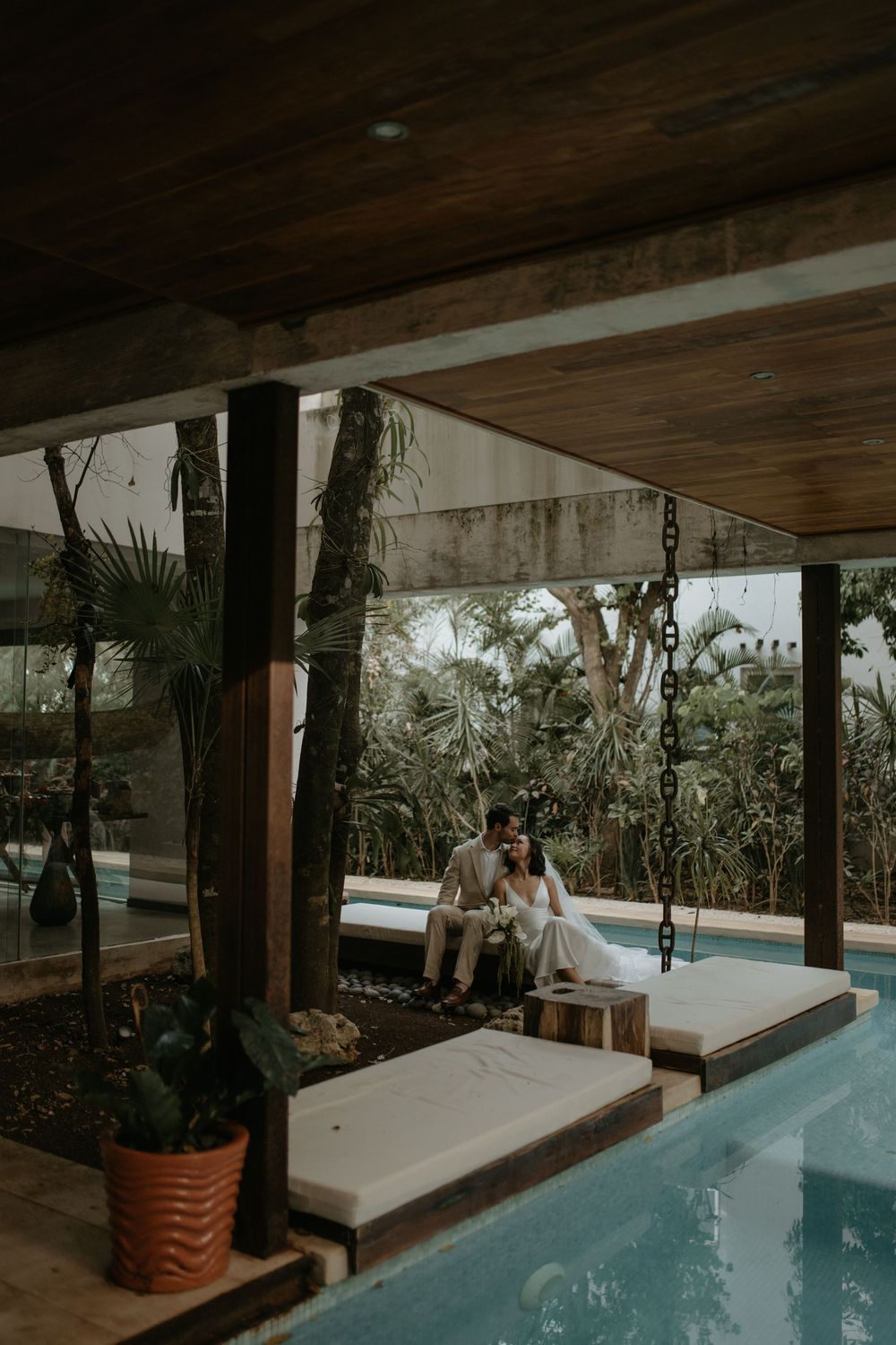 Couple in wedding attire sitting together on a poolside daybed surrounded by lush tropical greenery at a luxury villa.