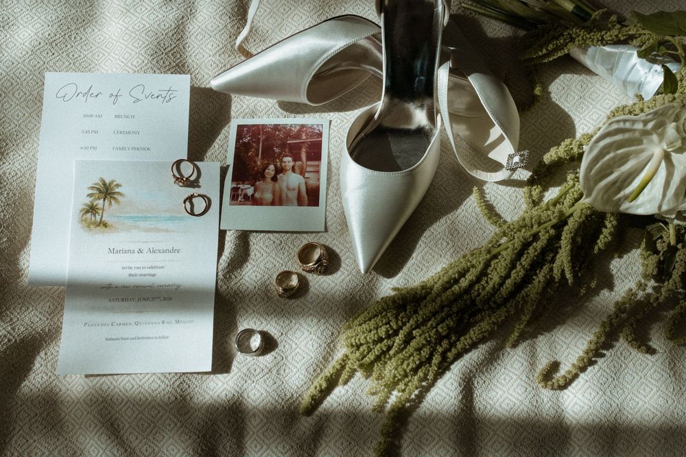 Elegant wedding flat lay with white heels, gold rings, order of service card, Polaroid photo, and dried greenery on white fabric.