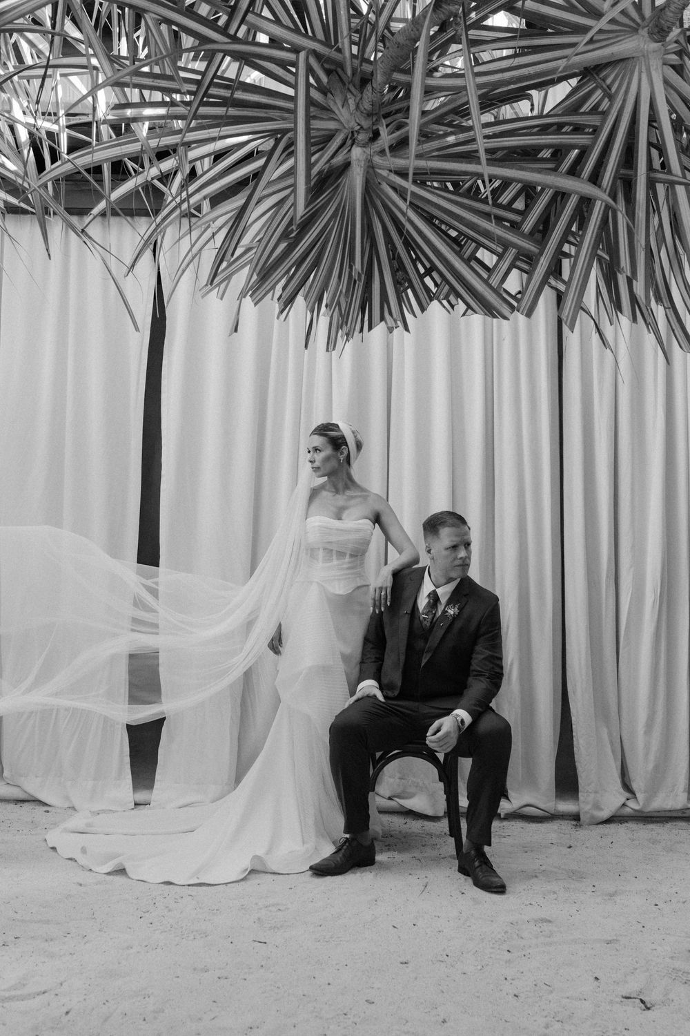 Black and white wedding photo of bride standing beside seated groom under palm fronds with flowing veil.