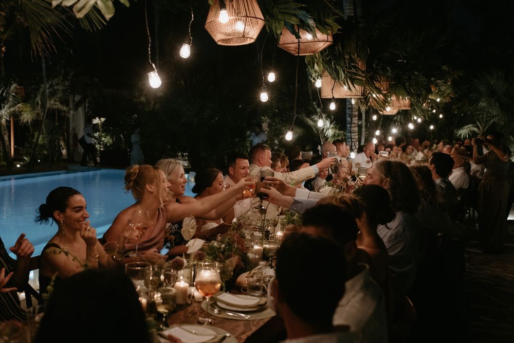 Guests raising glasses in a toast at an elegant outdoor wedding reception with string lights and tropical greenery at night.