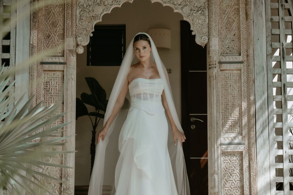 Elegant bride in strapless white wedding gown with veil standing in ornate doorway.