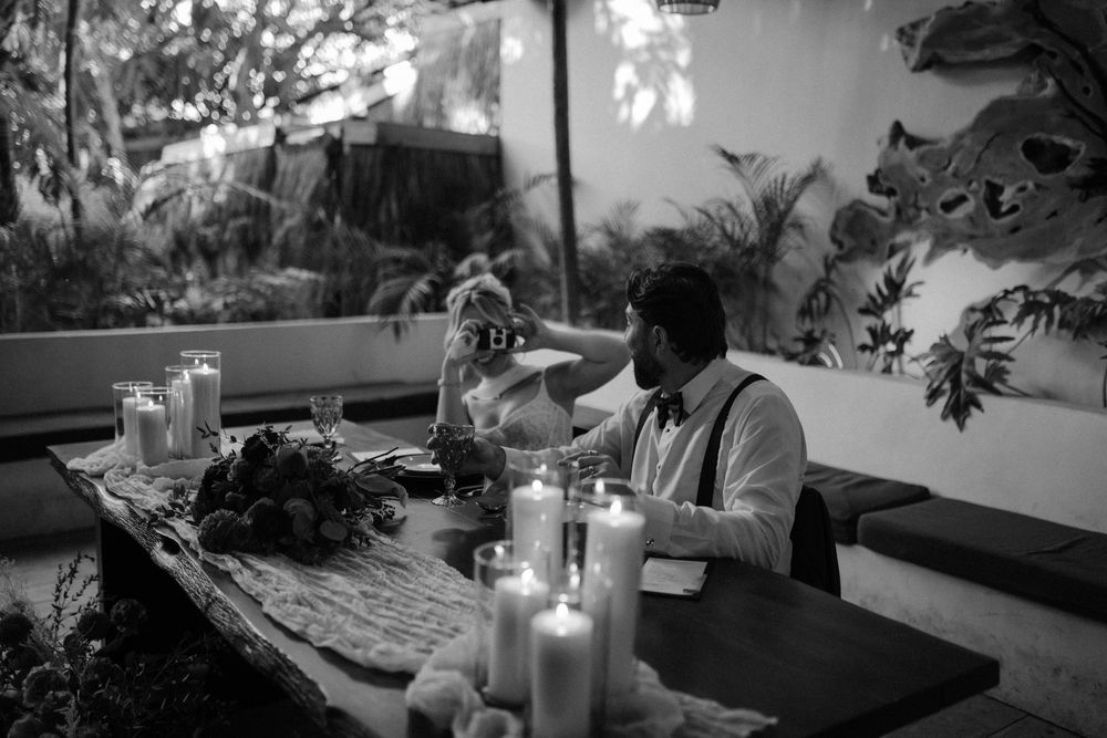 Black and white photo of bride and groom seated at candlelit table in lush tropical venue.