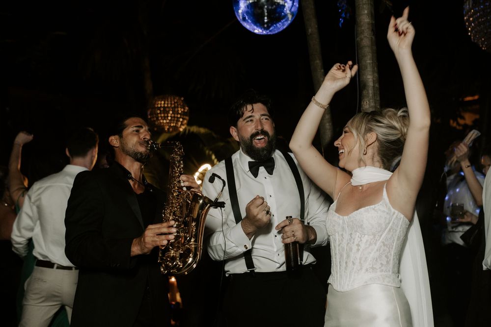 Bride dancing joyfully with saxophone player and guests at lively outdoor wedding reception.