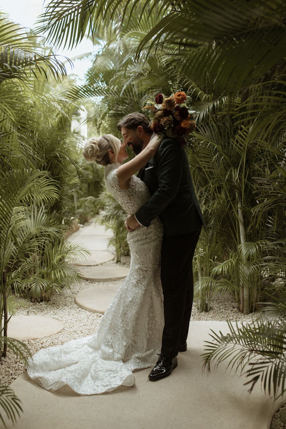 Bride in lace gown and groom in black suit embracing on tropical palm-lined garden path.