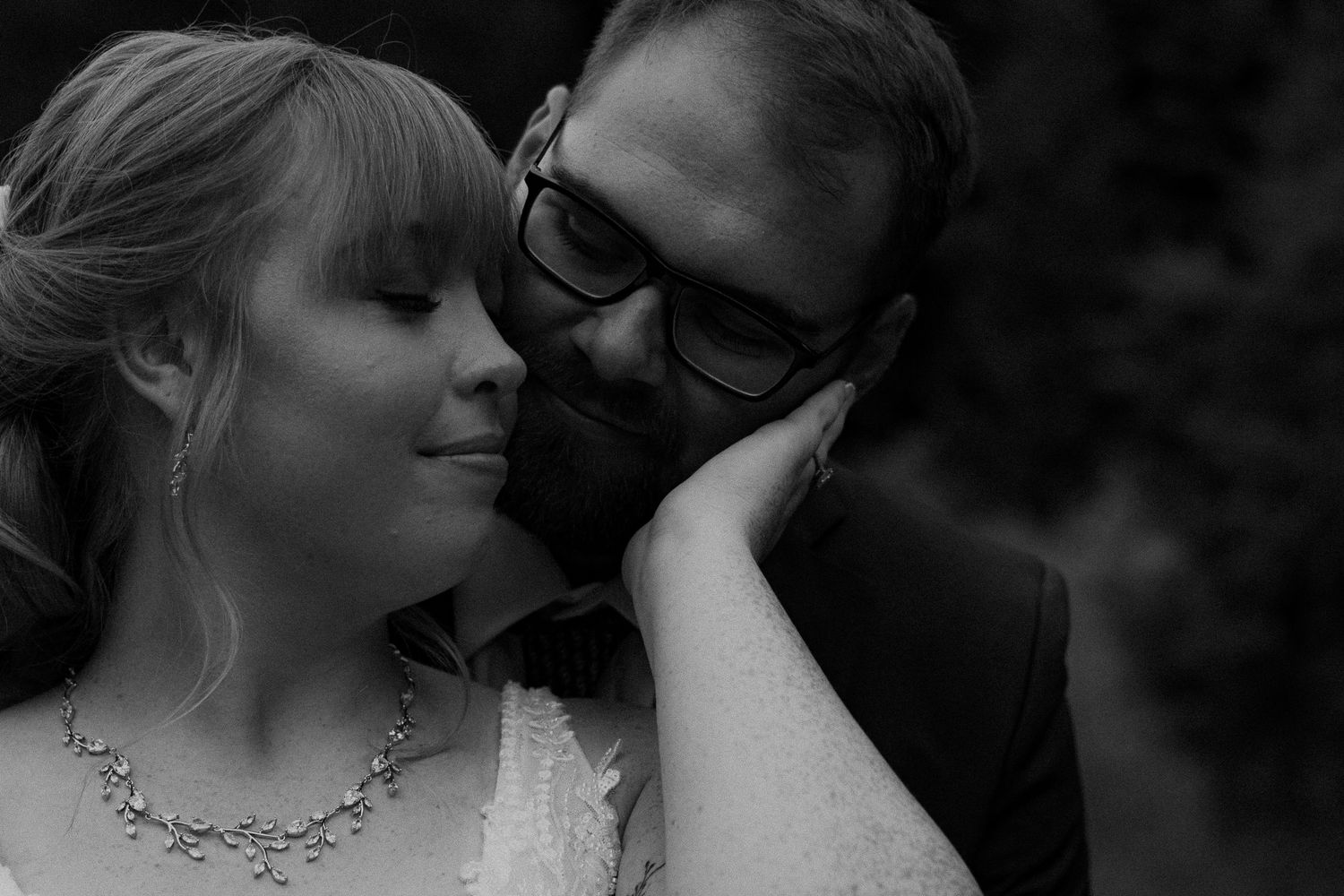 Bride in white lace gown and groom in navy suit share a romantic kiss outdoors in a forest setting.