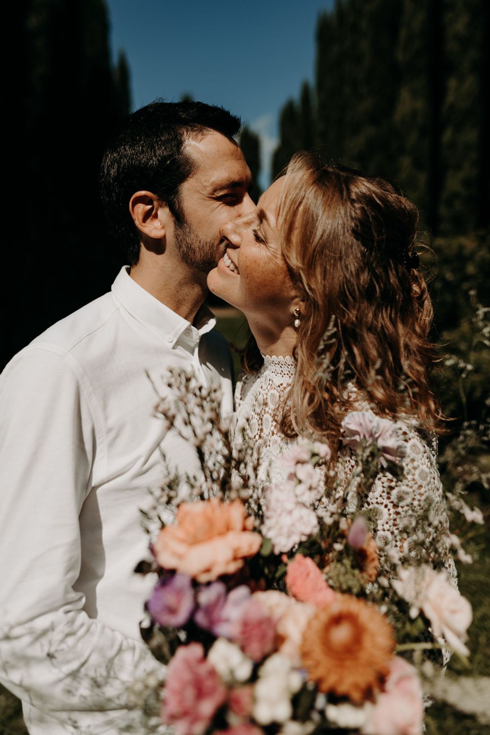 A romantic wedding couple shares a kiss outdoors while holding a bouquet of pink and peach flowers against cypress trees.