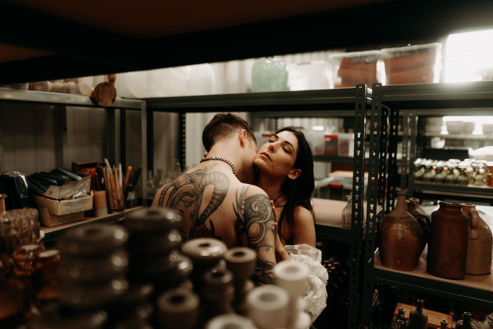 A romantic couple embraces in a dimly lit kitchen storage area among shelves of stored ingredients and supplies.