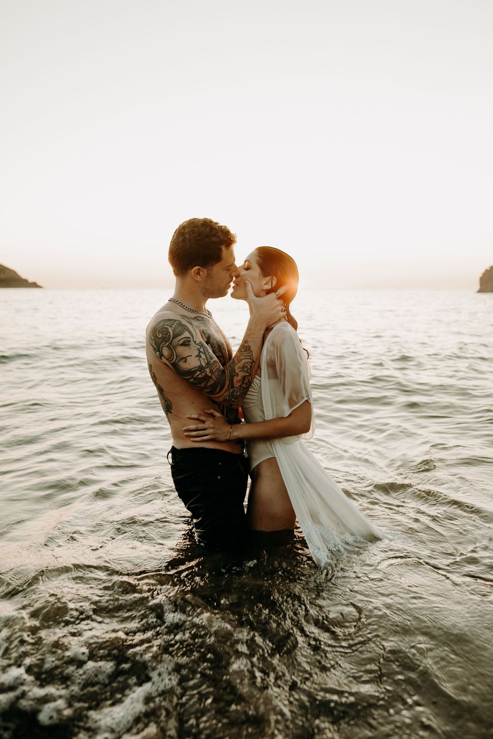 A romantic couple embraces in ocean waters at sunset against a misty coastal backdrop.