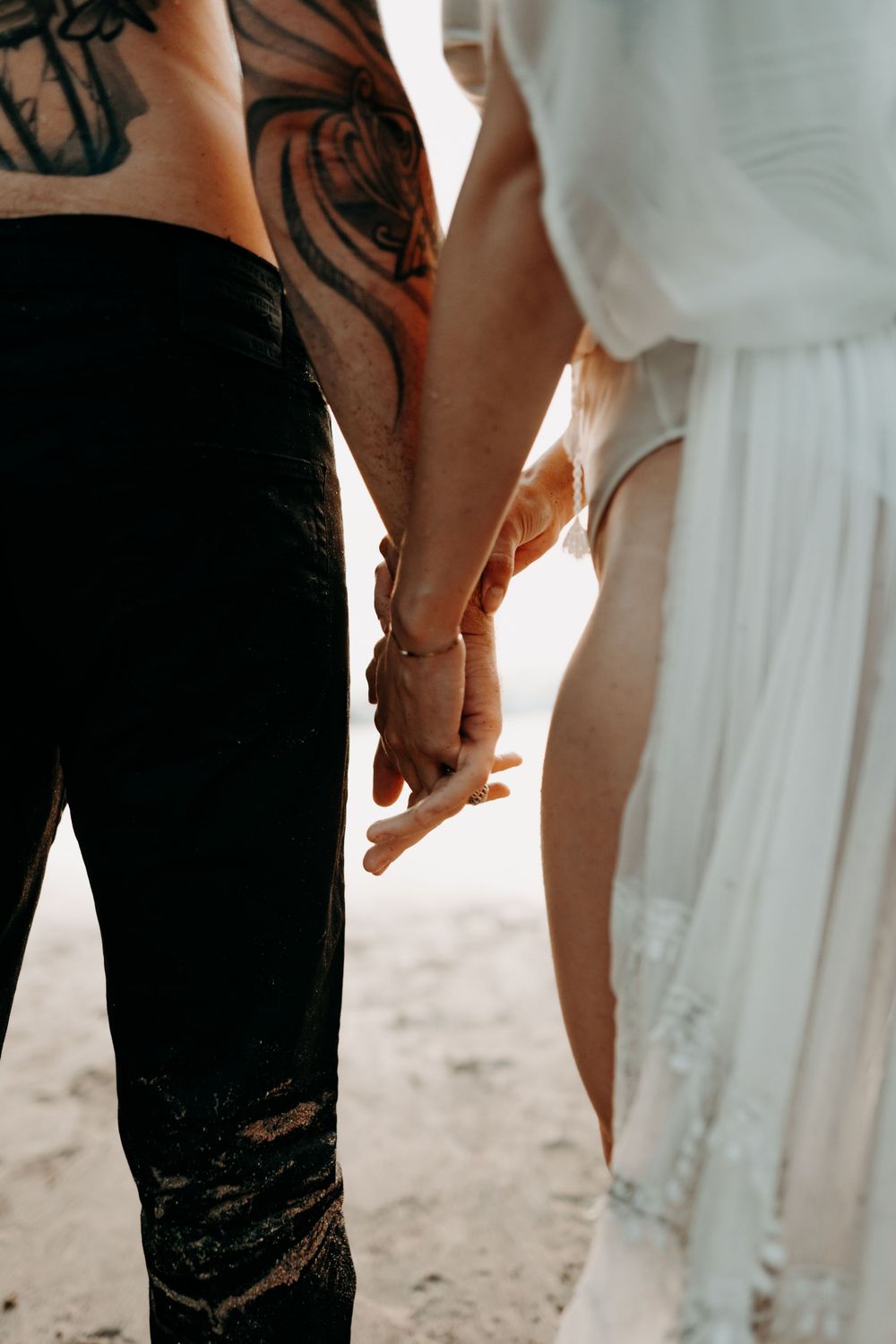A couple holding hands wearing contrasting black ripped jeans and a flowing white dress with tattoos visible.