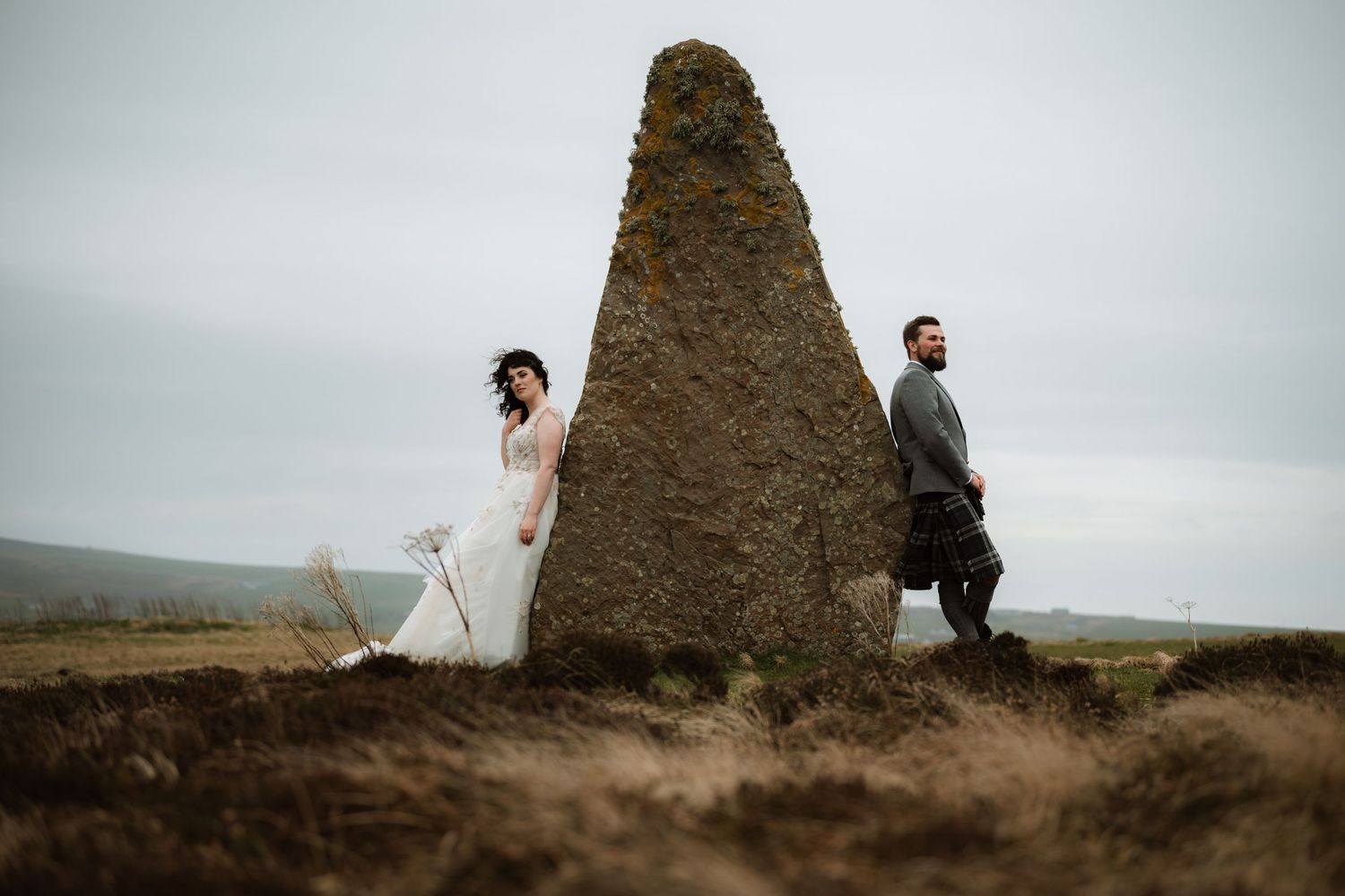 Newlyweds share a romantic kiss beneath a stone archway backlit by bright sunlight.