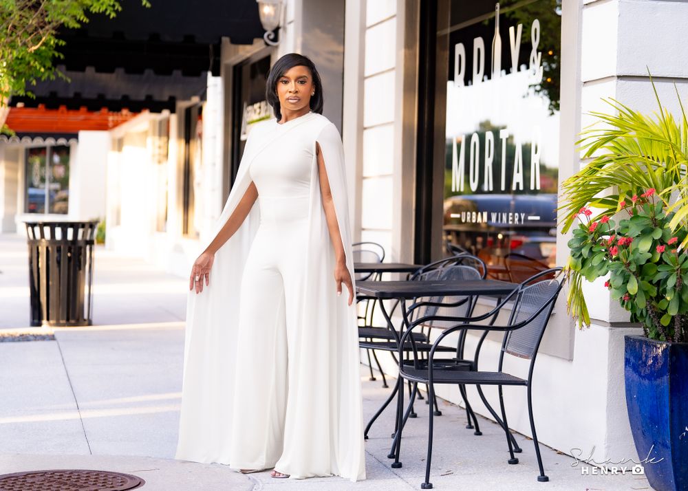 Elegant woman in white cape jumpsuit poses next to outdoor cafe seating with tropical plants.