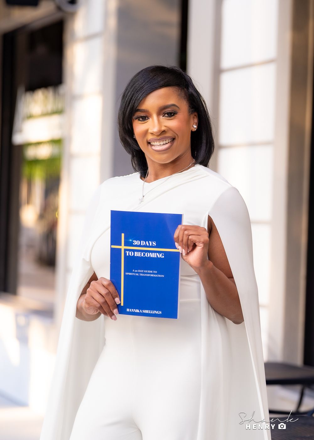 Woman in white outfit holds blue book while smiling outside building.