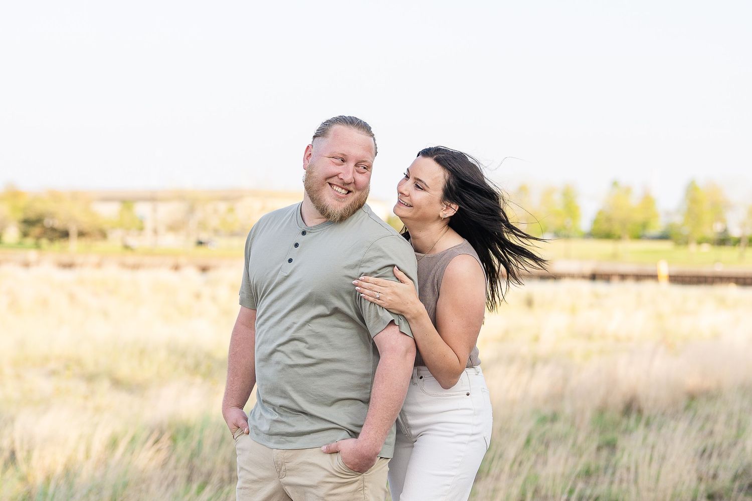 Smiling couple embracing in open field during golden hour, woman in white jeans and gray top.