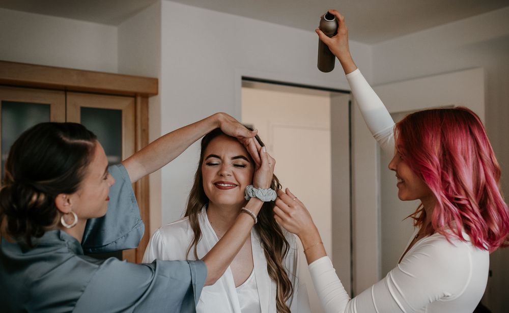 Two stylists helping a smiling bride with hair and makeup before her wedding.