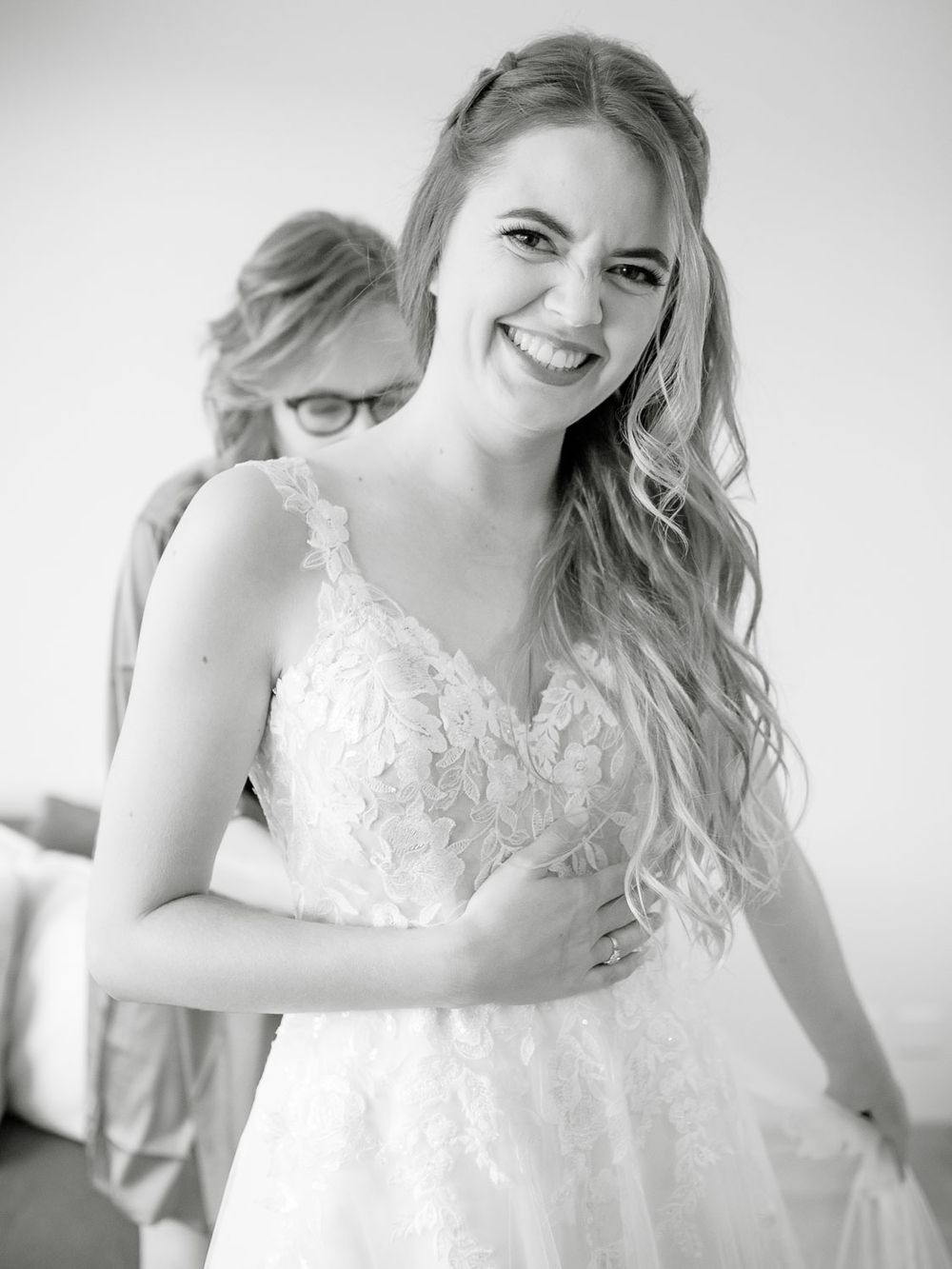 Black and white portrait of a smiling bride in lace gown being helped to dress.