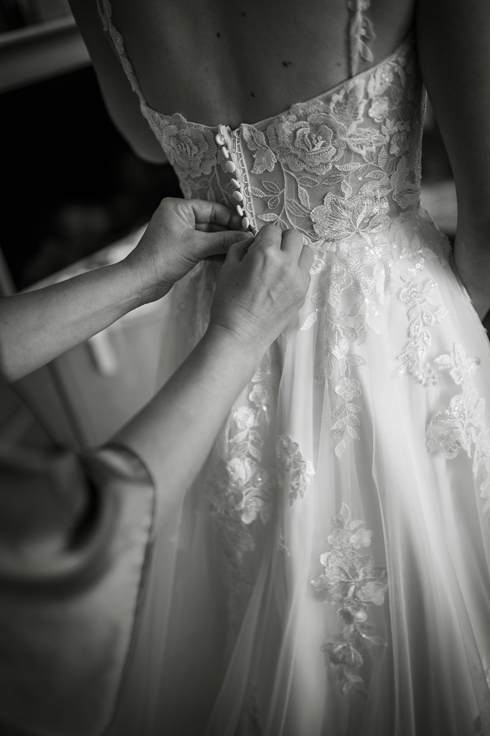 Black and white close-up of hands buttoning the back of a lace wedding gown.