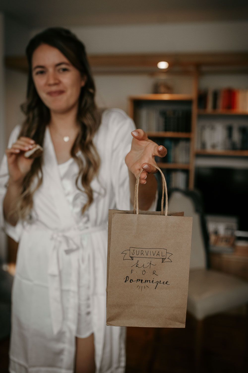Smiling bride in white robe holds a kraft paper survival kit gift bag personalized for Dominique.