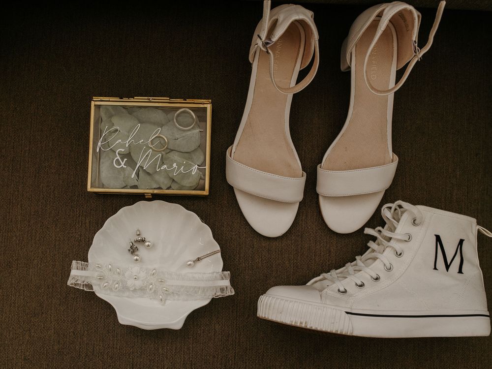 Flat lay of white wedding shoes, sneakers with monogram M, a ring box, and a lace garter on carpet.
