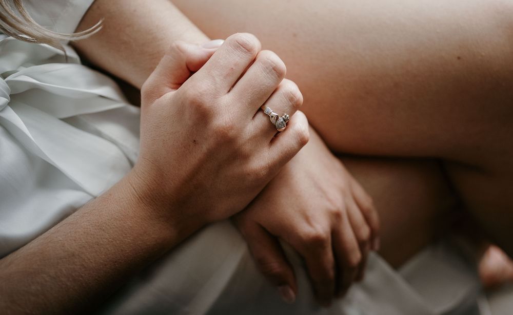 Close-up of bride's hands resting on her lap showcasing a sparkling diamond engagement ring against white fabric.