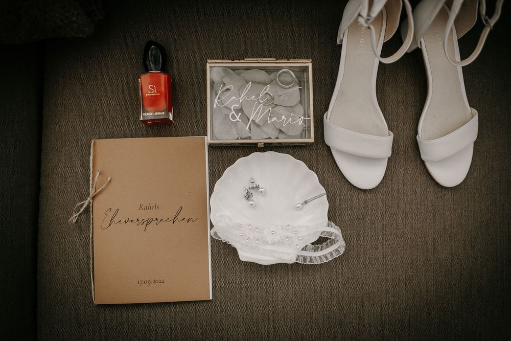 Bridal flat lay featuring white heeled sandals, jewelry dish, nail polish, and a vow book on a neutral surface.