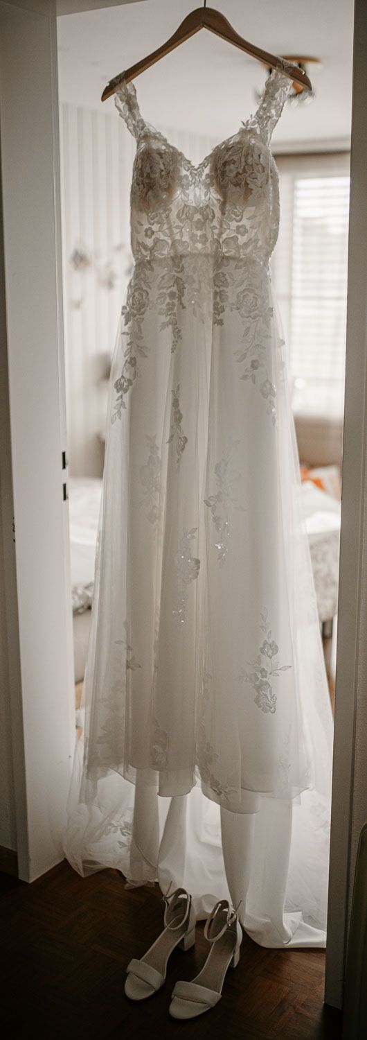 Elegant lace and tulle wedding dress hanging on a door with white strappy heels displayed below.
