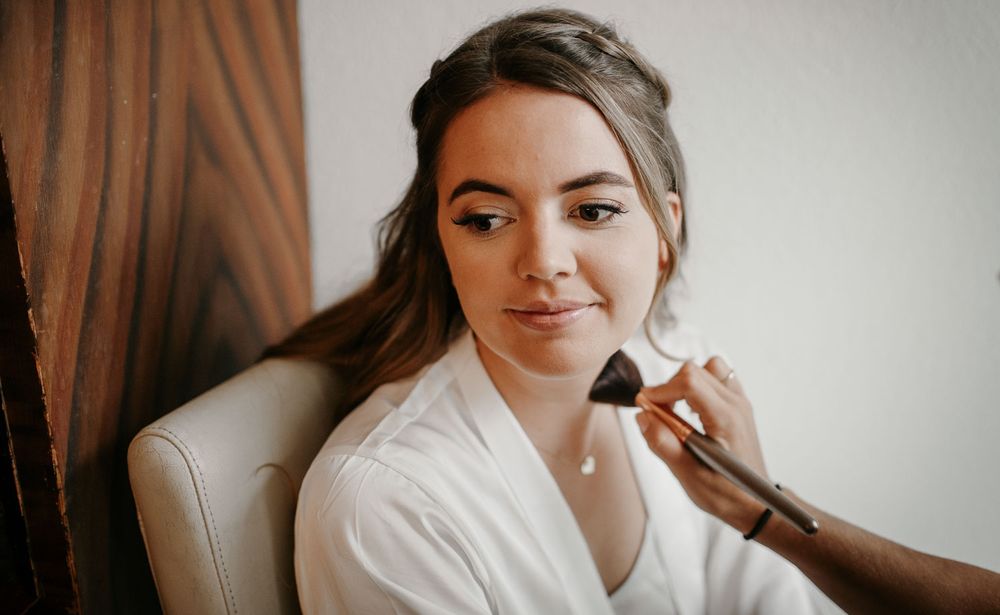 Beautiful bride in a white robe gets blush applied with a makeup brush during her wedding morning preparations.