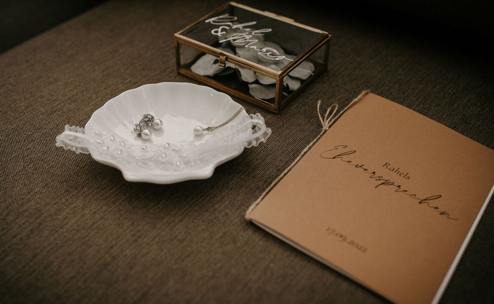 Wedding details flat lay with ring dish, jewelry, and personalized guest book on table.