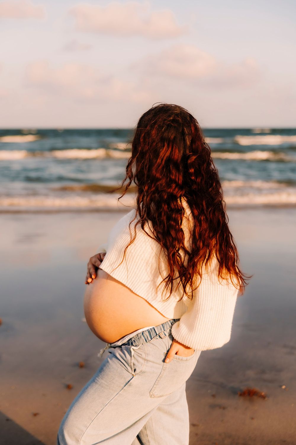 Pregnant woman in white sweater and jeans cradling baby bump on Galveston beach at golden hour taken by Houston Photographer.