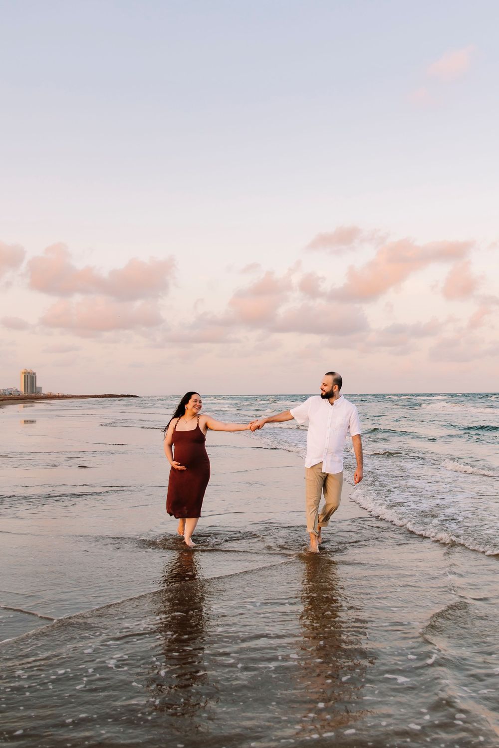 Pregnant couple holding hands walking barefoot on a sandy beach at sunset with gentle ocean waves in Galveston Texas by Houston Photographer