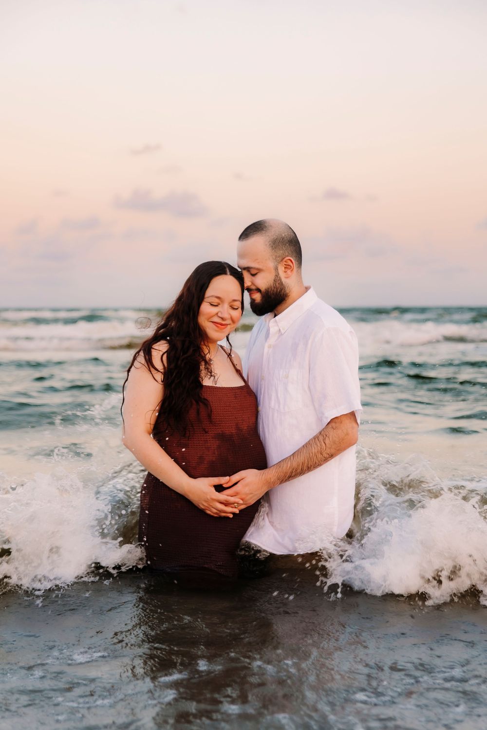 Expecting couple embraces tenderly in ocean waves at sunset, hands resting on pregnant belly taken by Houston Photographer.