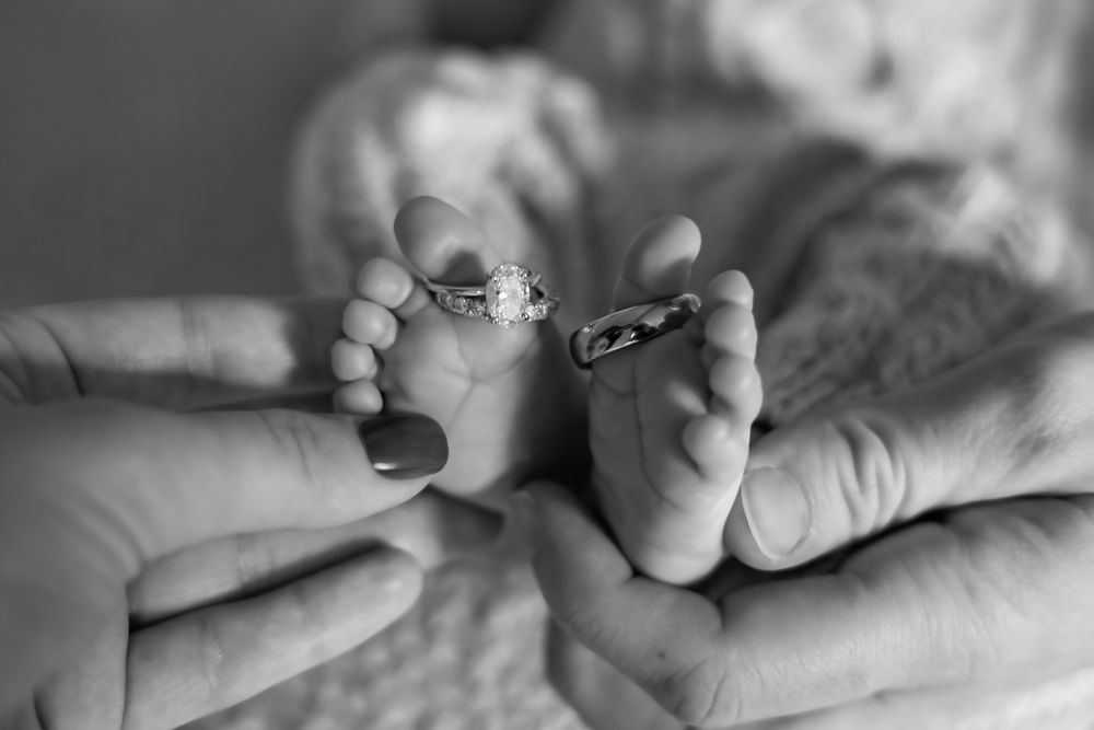 Black and white photo of newborn baby feet held by parents hands with wedding rings placed on tiny toes.
