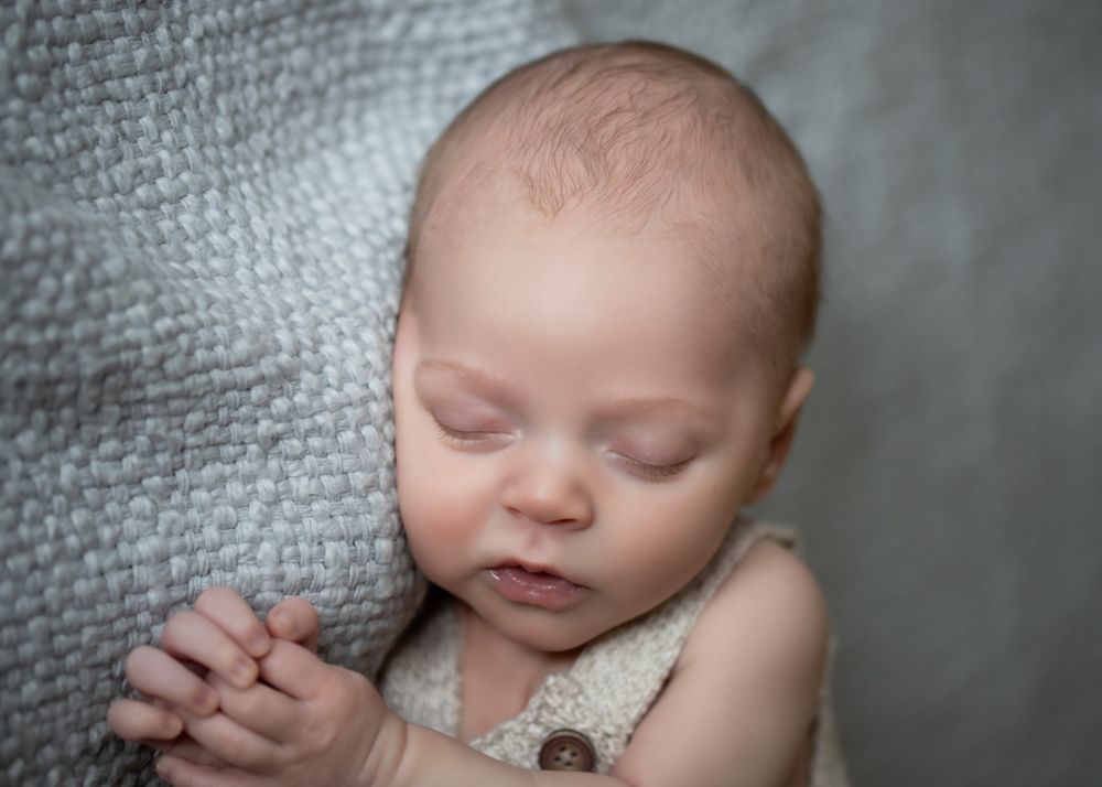 Peaceful newborn baby sleeping on a soft gray knit blanket, wearing a cream romper, in a professional portrait photo.