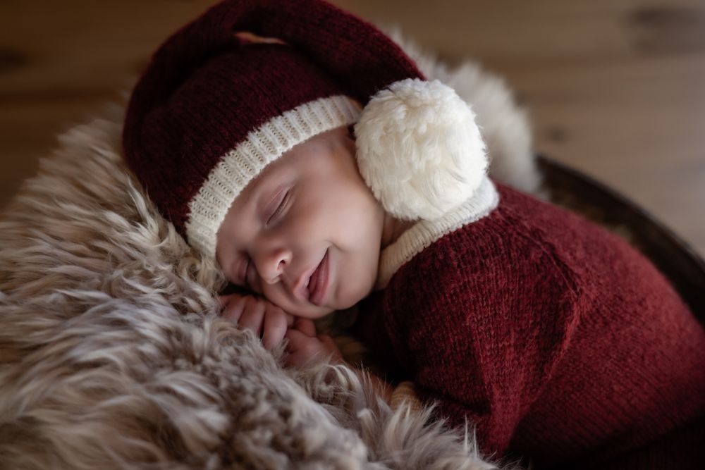 Sleeping newborn baby in red knit outfit and Santa hat with white pompom on fluffy fur blanket.