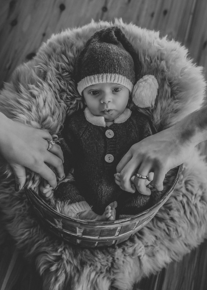 Black and white photo of a newborn baby in a knit hat lying in a fur-lined basket held by a parent.