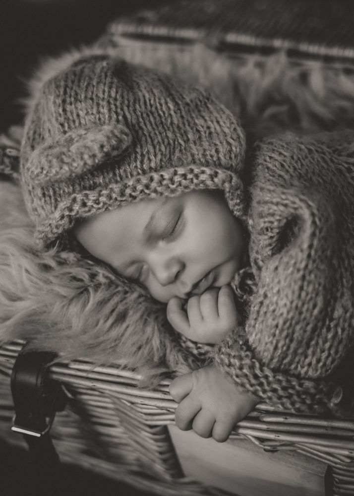 Black and white photo of a sleeping newborn baby in a knit hat, nestled in a wicker basket with fur.