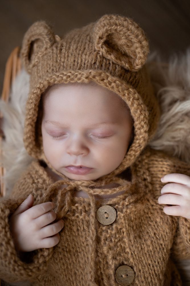 Sleeping newborn baby wearing a brown knit bear ear bonnet and matching sweater in a fur-lined basket.