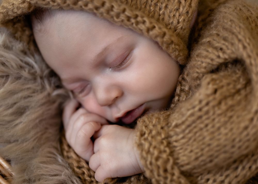 Peaceful newborn baby sleeping in brown knit outfit and bonnet, curled up on soft fluffy blanket.