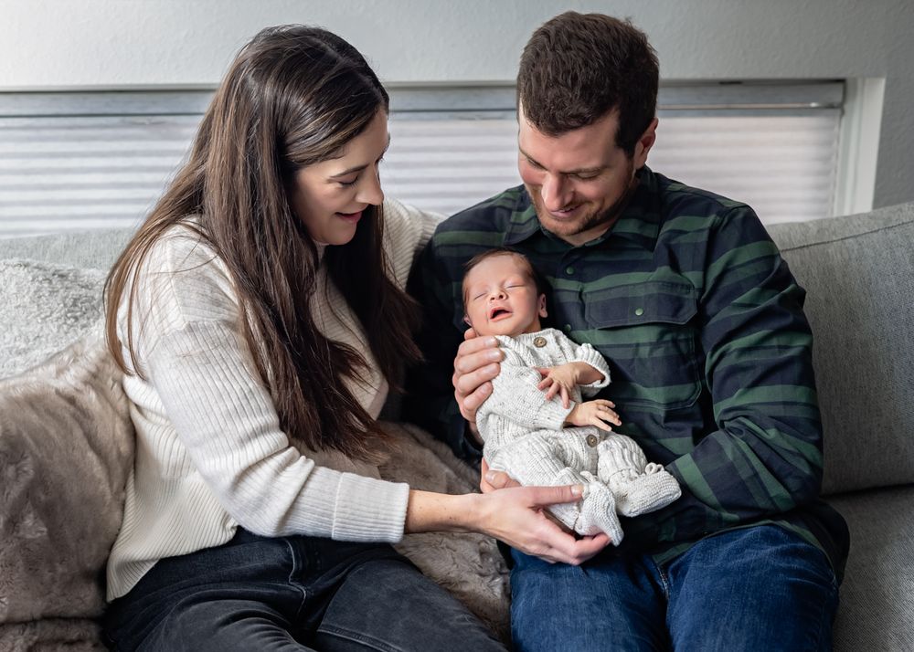 A young couple sitting on a couch smiling at their newborn baby wrapped in a white knit outfit.