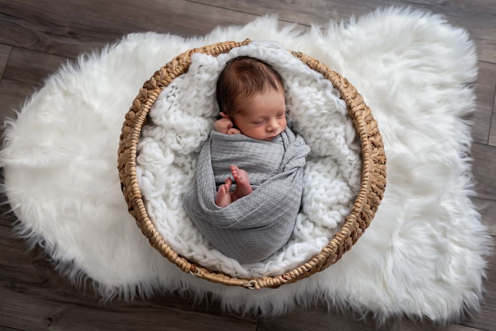 Newborn baby swaddled in gray wrap sleeping in wicker basket lined with white fur on wooden floor.