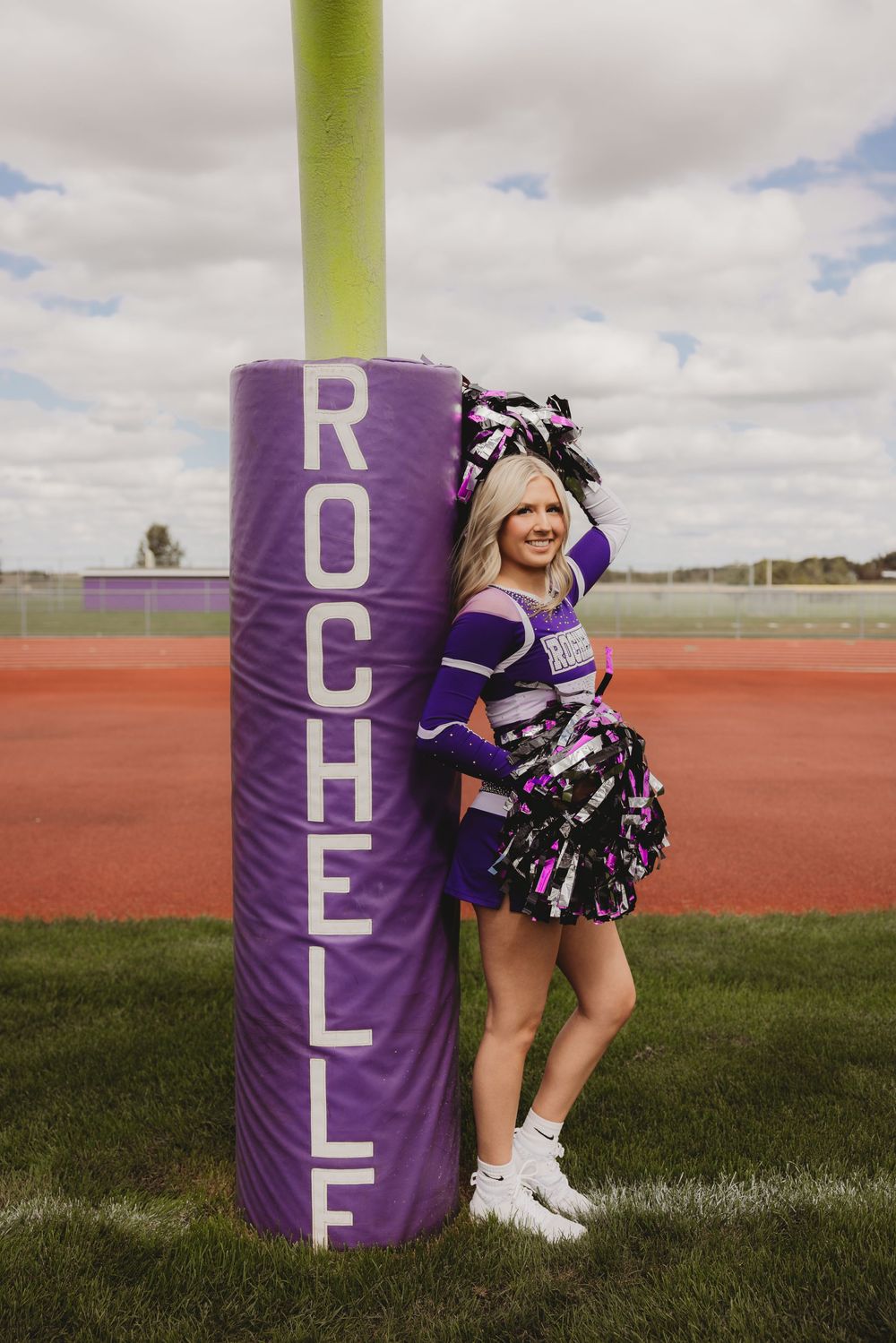 Rochelle High School cheerleader in purple uniform posing with pom-poms beside a purple goal post pad on a football field.