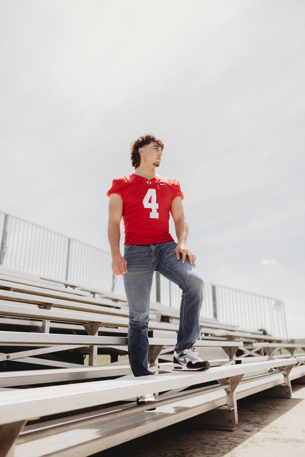 Young man in red football jersey number 4 and jeans standing on bleachers at an outdoor sports stadium.