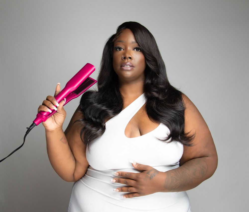 Plus size model in white dress holds pink hair styling tool against gray studio background.