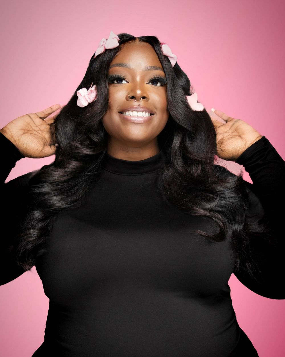 Person wearing a black sweater poses with flowing dark wavy hair adorned with small pink flowers against pink background.